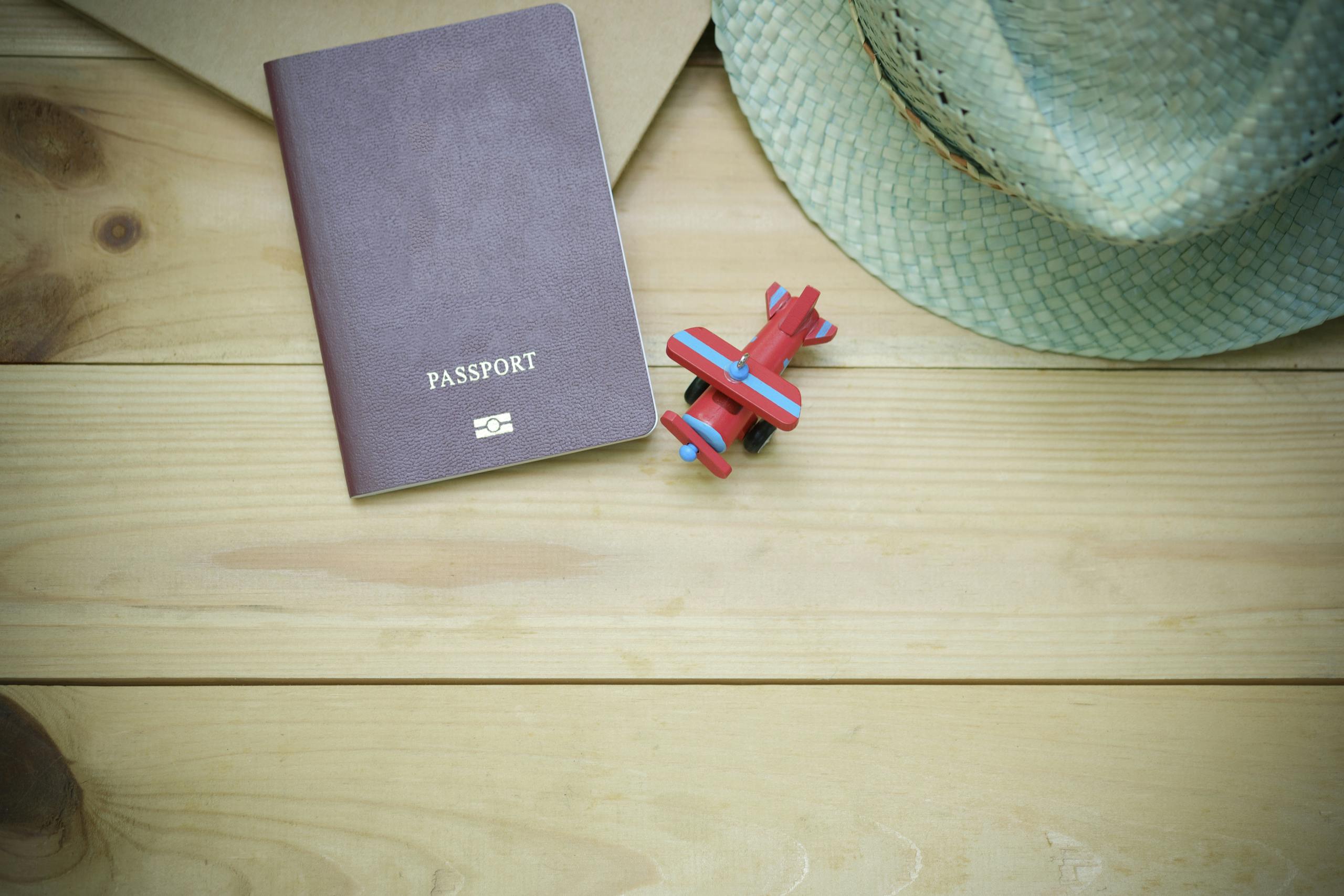 Flat lay of a passport, straw hat, and toy airplane on a wooden surface.